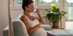 Pregnant woman drinking water on sofa at home mobile
